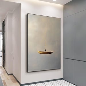Op de afbeelding: Een groot rechthoekig wandkunstwerk met een kleine gouden boot die zweeft in een uitgestrekte, wazige, lichtgrijze lucht. Een enkele dunne verticale gouden lijn strekt zich omhoog uit vanaf het midden van de boot, die een minuscuul donker silhouet draagt.