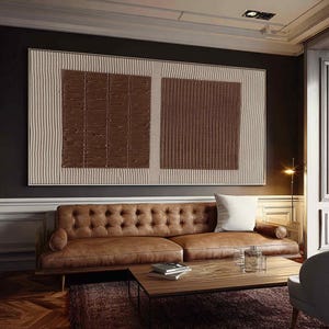 Puede incluir: Gran obra de arte mural abstracto con un dise&ntilde;o texturizado. La obra presenta dos cuadrados texturizados marrones sobre un fondo beige con rayas verticales. Un sof&aacute; de cuero marr&oacute;n y una mesa de centro de madera est&aacute;n frente a la obra.