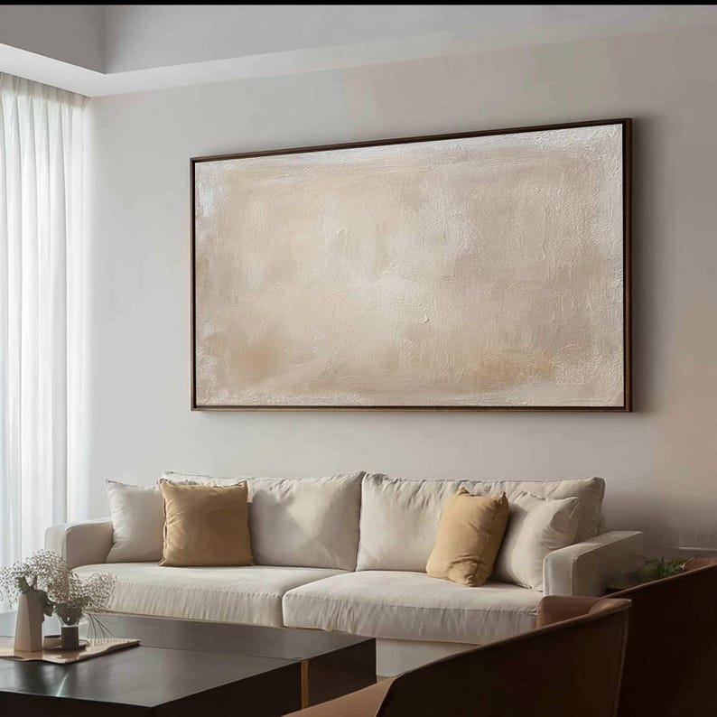 May include: A large, rectangular abstract painting with a textured, off-white surface, framed in dark brown. The artwork hangs above a cream-colored sofa with mustard-yellow throw pillows. A black coffee table and decorative items complete the living room setting.