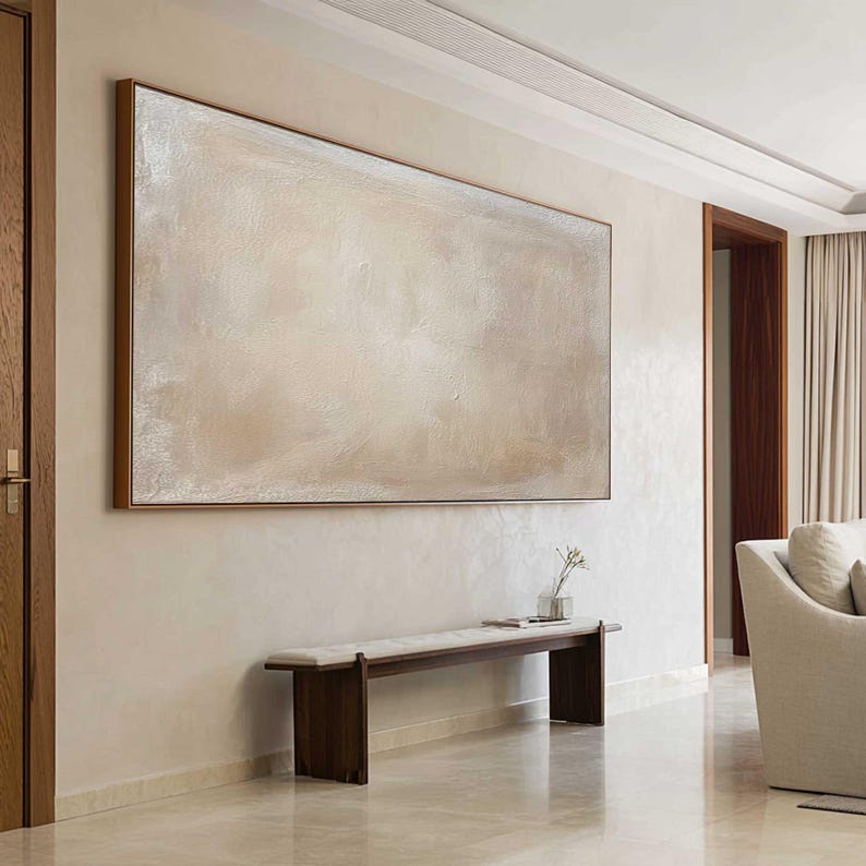 May include: A large, rectangular abstract painting in neutral tones hangs on a beige wall. The artwork is framed in a dark wood. Below the painting is a dark wood bench with a light-colored cushion. A vase with a small plant sits on the bench.