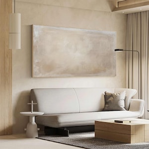May include: A large, rectangular abstract painting in neutral tones hangs above a white sofa. The artwork features a blend of beige and off-white hues. A wooden coffee table and a side table complete the modern living room setting.