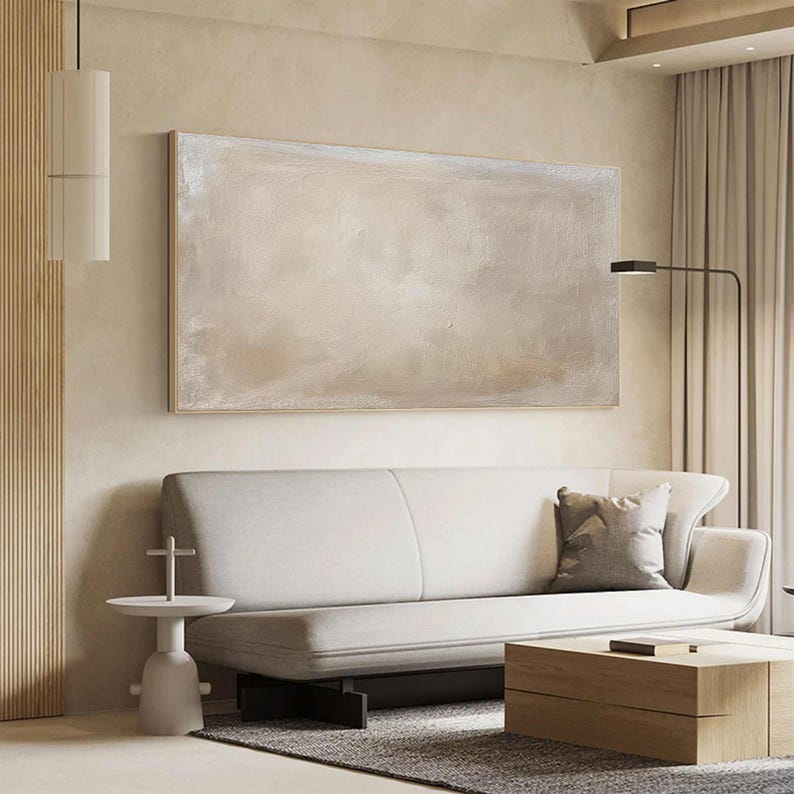 May include: A large, rectangular abstract painting in neutral tones hangs above a white sofa. The artwork features a blend of beige and off-white hues. A wooden coffee table and a side table complete the modern living room setting.