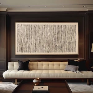May include: A large abstract artwork with vertical black lines on a cream background hangs above a cream-colored tufted sofa. The sofa has black velvet pillows and a patterned pillow. A dark wood coffee table and a decorative vase are in front of the sofa.
