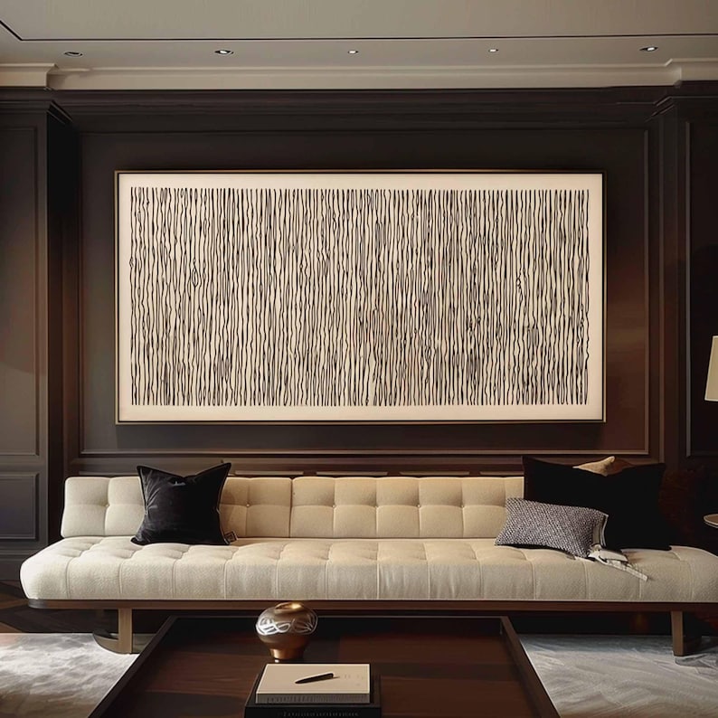 May include: A large abstract artwork with vertical black lines on a cream background hangs above a cream-colored tufted sofa. The sofa has black velvet pillows and a patterned pillow. A dark wood coffee table and a decorative vase are in front of the sofa.