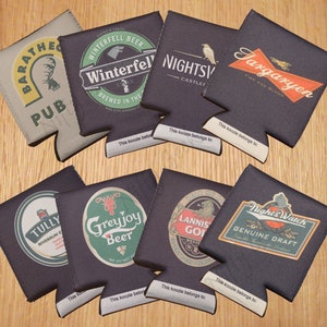 May include: Ten different koozies with various designs and text. The koozies are all black, brown, or gray with white text. The text includes the names of houses from the TV show Game of Thrones, such as Targaryen, Winterfell, and Greyjoy.