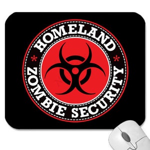 以下が含まれることがあります： 中央に白い円形がある黒と赤の丸いマウスパッド。円形には黒いバイオハザードシンボルと、外側に「Homeland Zombie Security」という文字が書かれている。