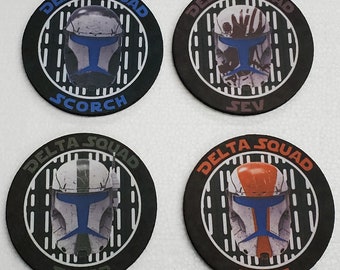 May include: Four round black coasters with white and gray stripes. Each coaster features a different Star Wars Clone Trooper helmet with a blue visor. The text "DELTA SQUAD" is printed in white on each coaster. The names "SCORCH", "SEV", "FIXER", and "BOSS" are printed in white on each coaster.