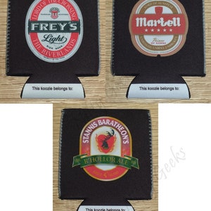 May include: Three black and white can koozies with different designs. The first koozie features a circular logo with the text "Frey's Light" and "The Riverlands". The second koozie features a circular logo with the text "Martell" and "Prince of Dorne". The third koozie features a circular logo with the text "Stannis Baratheon's" and "R'hollor Ale".
