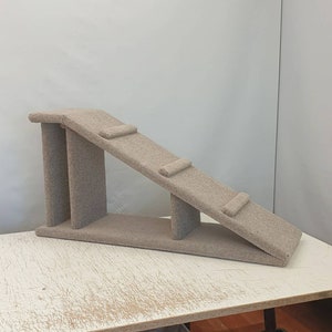 Puede incluir: Una rampa beige para gatos con superficie alfombrada. La rampa tiene una plataforma en la parte superior, una secci&oacute;n inclinada con dos peque&ntilde;os escalones y una base. La estructura est&aacute; dise&ntilde;ada para ayudar a los gatos a trepar.