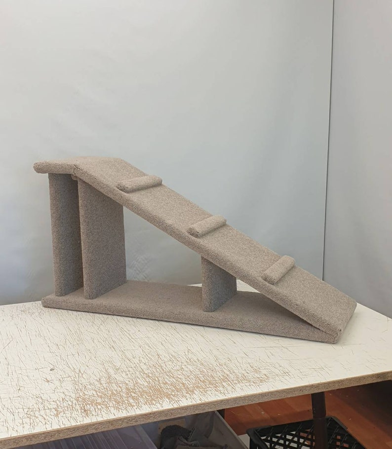 Puede incluir: Una rampa beige para gatos con superficie alfombrada. La rampa tiene una plataforma en la parte superior, una secci&oacute;n inclinada con dos peque&ntilde;os escalones y una base. La estructura est&aacute; dise&ntilde;ada para ayudar a los gatos a trepar.