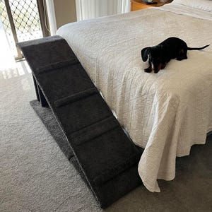 Puede incluir: Una rampa gris oscuro para mascotas con tres escalones se coloca junto a una cama con un edred&oacute;n blanco acolchado. Un peque&ntilde;o perro salchicha negro y marr&oacute;n est&aacute; en la cama. La rampa est&aacute; dise&ntilde;ada para ayudar a las mascotas a acceder a la cama.
