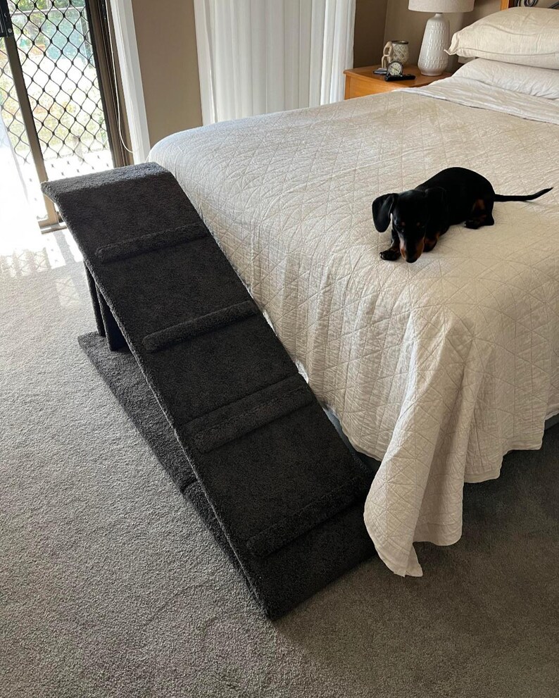 Puede incluir: Una rampa gris oscuro para mascotas con tres escalones se coloca junto a una cama con un edred&oacute;n blanco acolchado. Un peque&ntilde;o perro salchicha negro y marr&oacute;n est&aacute; en la cama. La rampa est&aacute; dise&ntilde;ada para ayudar a las mascotas a acceder a la cama.