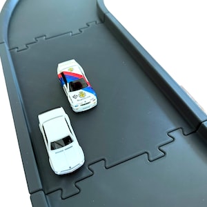 May include: Two toy cars on a dark grey modular plastic track. One car is white with red, blue, and yellow racing decals and the text Warsteiner 40. The other car is solid white. The track pieces feature interlocking puzzle-style edges.