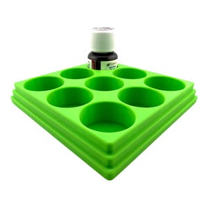 Può includere: Un vassoio organizzatore per vernici quadrato in plastica verde brillante con nove alloggiamenti circolari incassati per contenere piccoli flaconi. Un singolo flacone con tappo bianco ed etichetta scura è posizionato nello slot centrale.