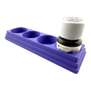 May include: A purple plastic paint bottle holder with three empty circular slots and one slot containing a Humbrol Dark Green Wash bottle with a white ribbed cap. The rectangular stand features a stepped design with visible layer lines.