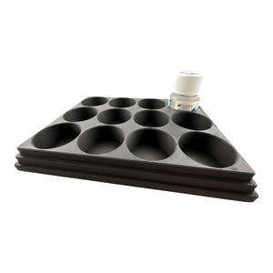 May include: A dark gray, triangular paint organizer tray featuring twelve circular recessed wells of varying sizes arranged in rows. A small, cylindrical white bottle with a screw-top lid and a label is positioned in the back corner of the tray.