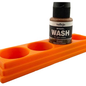 May include: An orange plastic paint holder with three circular wells and a stepped design. A bottle of Vallejo Model Wash, for light colors, sits in one well. The bottle is brown with a black cap.