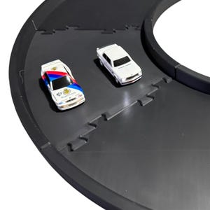 May include: Two miniature toy cars on a curved black plastic track. One car is white with blue and red racing stripes and Warsteiner branding, while the other is a solid white car. The track segments feature interlocking puzzle-like edges.
