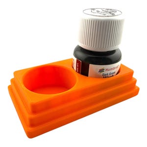 May include: A bright orange plastic stand with two circular recessed holders, one of which contains a small bottle of Humbrol Dark Green Wash with a white ribbed cap. The stand features a stepped, tiered design with a rectangular base structure.