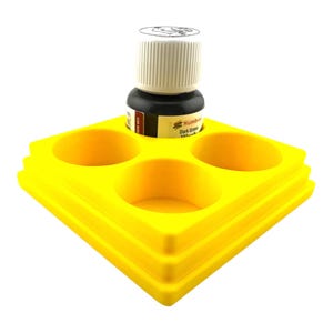 May include: A bright yellow square tiered stand with three circular indentations for holding small containers. A small bottle of Humbrol Dark Green Wash with a white ribbed cap is placed inside one of the circular openings on the yellow stand.