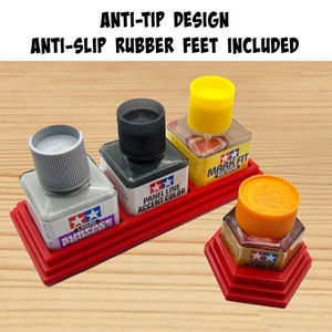 May include: Red plastic organiser with four slots for holding small bottles of paint. The organiser has a non-slip rubber base and an anti-tip design. The bottles are labelled "Tamiya Liquid Surface Primer", "Tamiya Panel Line Accent Color", "Tamiya Mark Fit Decal Solution", and "Tamiya Cement".