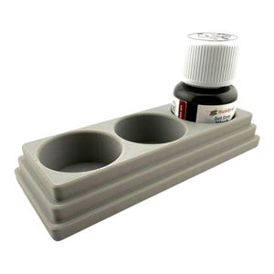 May include: A gray 3D-printed rectangular paint bottle holder with two circular recessed slots. One slot contains a small Humbrol Dark Green Wash paint bottle with a white cap and a label featuring the brand name and product details.