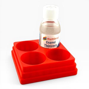 May include: A bottle of Humbrol Enamel Thinners with a white cap stands on a red paint tray. The tray has four circular wells. The label on the bottle is visible. The product is for thinning enamel paints.