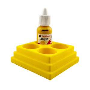 May include: A yellow paint bottle with a white cap sits in a yellow tiered paint holder. The paint bottle is labelled "Humbrol Acrylic" and has the number "69". The paint holder has four circular openings for paint bottles.