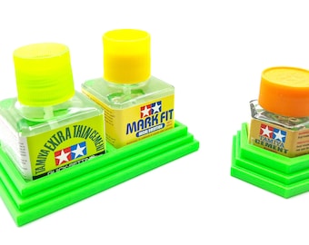 Op de afbeelding: Drie flesjes Tamiya modellijm op een groene plastic standaard. De flesjes zijn gelabeld met "Tamiya Extra Thin Cement", "Tamiya Mark Fit Decal Solution" en "Tamiya Cement".