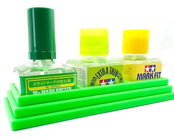 Op de afbeelding: Drie flessen modelbouwvloeistoffen in een groene kunststofhouder. De flessen zijn gelabeld met "Mr. Hobby Mark Softer", "Miwa Extra Thin Cement" en "Mark Fit".