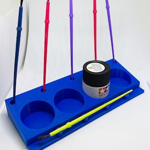 May include: Blue plastic paint brush holder with five slots for brushes and a small container for paint. The holder has three round depressions for paint pots. A yellow brush and a bottle of Tamiya Acrylic paint are visible.