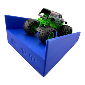 Könnte beinhalten: Ein blauer, abgewinkelter Präsentationsständer mit dem Text "JEFF DID IT!" hält ein grünes und schwarzes Grave Digger Monster Truck Spielzeug. Der Spielzeug-Truck hat große schwarze Reifen und eine detaillierte Karosserie.