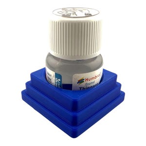 May include: A small bottle of Humbrol Enamel Thinners with a white ribbed cap, featuring a clear glass body and a white label with blue text. The bottle is securely held within a square, tiered blue plastic stand with a central circular cutout.