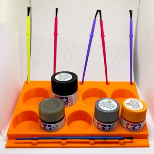 May include: Orange paint holder with six paint pots and five paint brushes. The paint pots are labeled "X-1", "X-10", "X-11" and "X-19". The paint brushes are yellow, pink, purple, and blue.