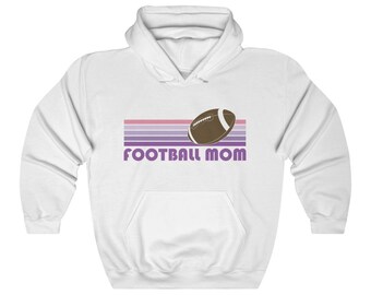 Może przedstawiać: Biała bluza z kapturem z paskami w kolorze fioletowym i różowym oraz grafiką brązowego futbolu. Tekst "FOOTBALL MOM" jest nadrukowany na fioletowo pod grafiką.