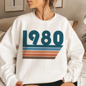 May include: White sweatshirt with a blue and brown graphic design featuring the year 1980 and colorful stripes.