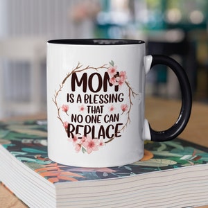 Puede incluir: Taza de cerámica blanca con interior y asa negros. La taza presenta el texto "MOM is a blessing that no one can replace" rodeado de un diseño de corona floral. La taza está sobre un libro con una cubierta floral.