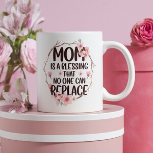 May include: White ceramic mug with the text "MOM is a blessing that no one can replace" surrounded by a floral wreath. The mug is on a pink round box with a white rim, with pink flowers and a rose in the background.