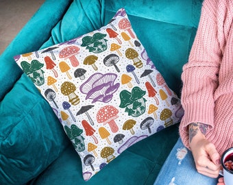 May include: Square cushion featuring a repeating pattern of colourful mushrooms in red, green, yellow, brown, and purple on a white background. The cushion is on a teal green sofa.
