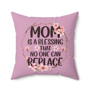 Puede incluir: Cojín cuadrado lila con el texto "MOM IS A BLESSING THAT NO ONE CAN REPLACE" en escritura negra. El texto está rodeado por una corona de ramas marrones y flores rosas. Un artículo decorativo.