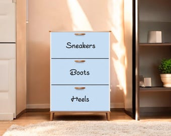 May include: A light blue shoe storage cabinet with three drawers labeled "Sneakers", "Boots", and "Heels" in black script. The cabinet has wooden handles and legs, and sits on a light-colored rug.