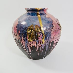 May include: A decorative ceramic vase with a rounded form. The vase displays a colour palette of pink, blue, and black with gold accents. The design includes a textured surface and a dripping paint effect, enhancing its artistic appeal.