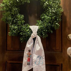 Puede incluir: Una corona verde y frondosa cuelga de una puerta de madera. Una faja de tela blanca está atada a la corona, con un cascanueces bordado y la letra "B" en azul claro. La puerta tiene un acabado marrón oscuro.