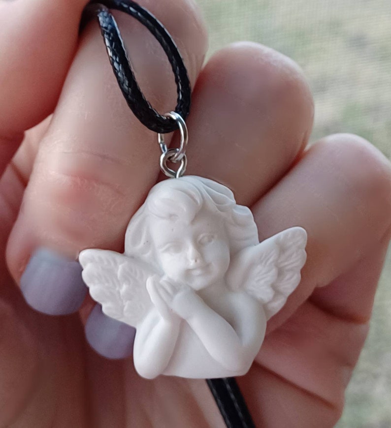 Może przedstawiać: Biały naszyjnik z zawieszką w kształcie cherubina z czarnym sznurkiem. Cherubin ma złożone dłonie do modlitwy i szczeg&oacute;łowe skrzydła. Zawieszka jest przymocowana do sznurka srebrnym pierścieniem.