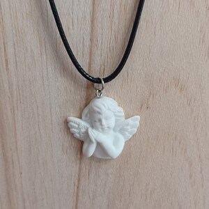 Może przedstawiać: Naszyjnik z białą zawieszką w kształcie cherubina na czarnym sznurku. Cherubin jest w pozycji modlitewnej ze szczeg&oacute;łowymi skrzydłami i kręconymi włosami. Zawieszka jest przymocowana do srebrnego uchwytu.