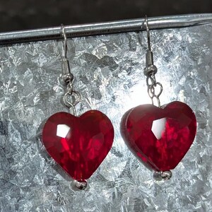 May include: A pair of red heart-shaped earrings with silver hooks. The earrings are made of faceted glass and have a shiny finish.
