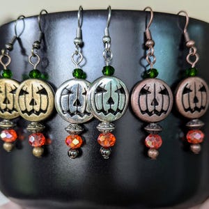 May include: Four pairs of dangle earrings with a jack-o'-lantern charm, green beads, and orange beads. The jack-o'-lantern charms are silver, gold, and copper.