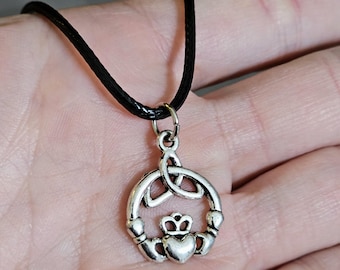 Pode incluir: Um colar de prata com um nó celta em um cordão de couro preto. O pingente apresenta um coração com uma coroa no topo, cercado por um círculo de nós entrelaçados.