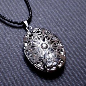 May include: Silver-toned oval pendant necklace with intricate openwork design. The pendant hangs from a black cord. The filigree pattern features floral and geometric shapes, with a central crystal accent.