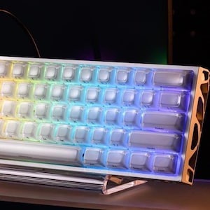 May include: A white mechanical keyboard with rainbow LED backlighting. The keyboard has a silver frame and clear keycaps. The keys are arranged in a compact layout. The lighting transitions across the keys, creating a vibrant effect.